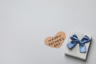 Heart shaped card with phrase HAPPY FATHER'S DAY and gift box on light grey background, flat lay. Space for text