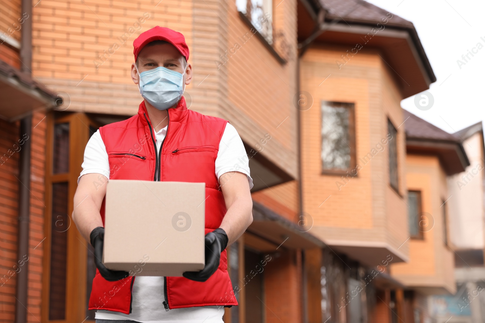 Photo of Courier in protective mask and gloves with box near house outdoors. Delivery service during coronavirus quarantine