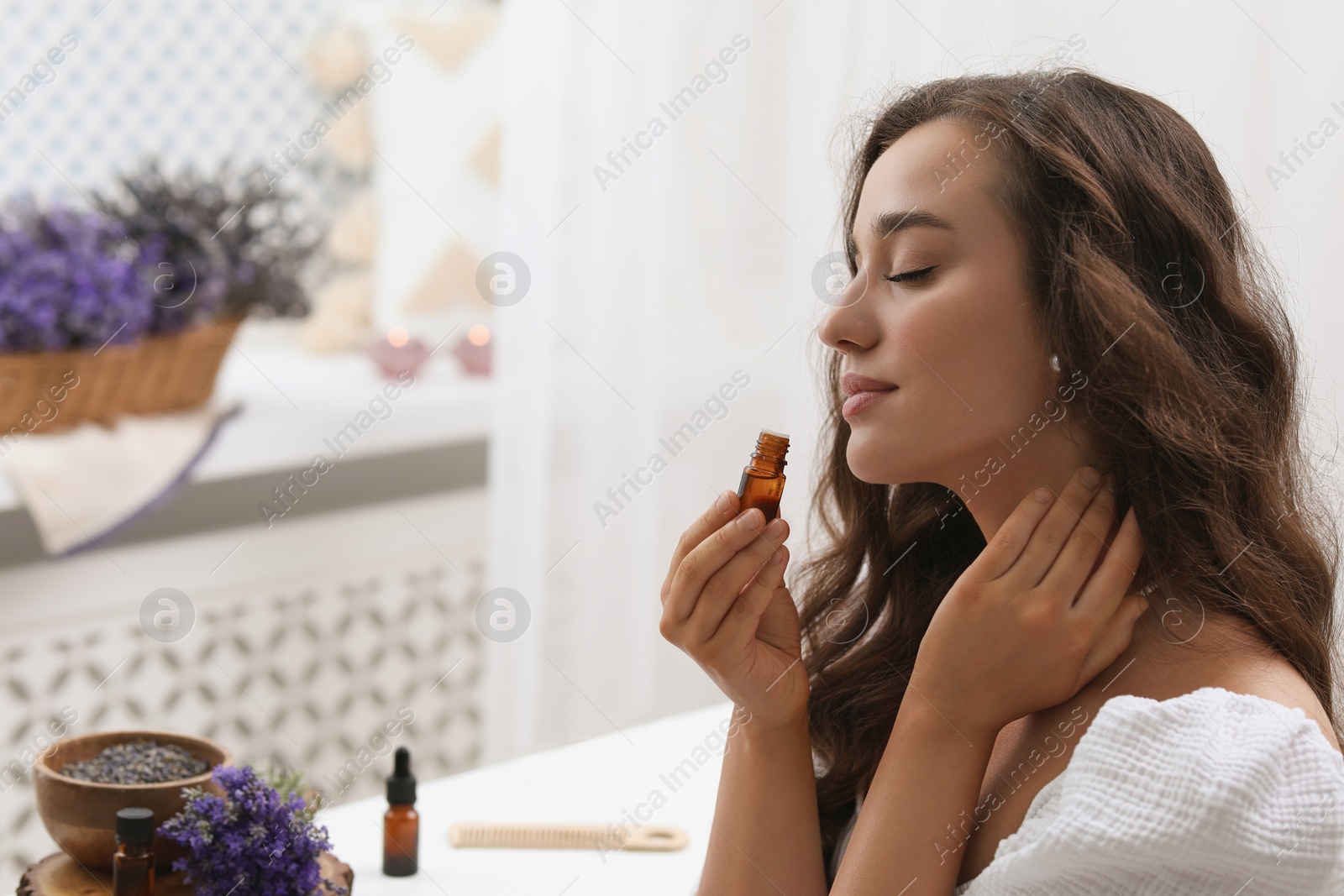 Photo of Beautiful young woman applying essential oil onto neck indoors