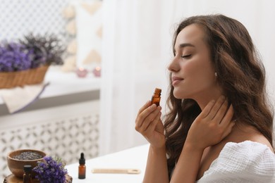 Beautiful young woman applying essential oil onto neck indoors