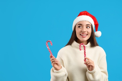 Young woman in beige sweater and Santa hat holding candy canes on blue background, space for text