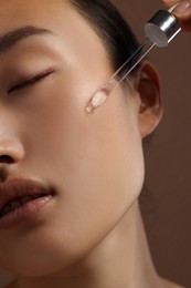 Beautiful young woman applying cosmetic serum onto her face on brown background, closeup