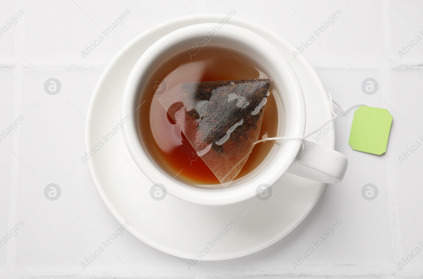 Photo of Tea bag in cup with hot drink on white tiled table, top view