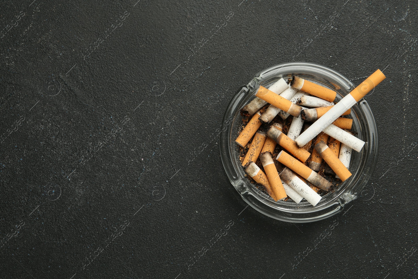 Photo of Glass ashtray full of cigarette stubs on black table, top view. Space for text