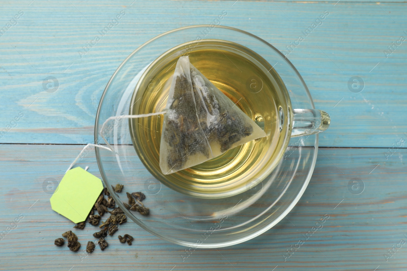 Photo of Tea bag in cup with hot drink and dry leaves on light blue wooden table, top view