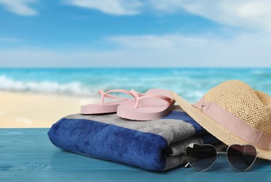 Beach towel, flip flops, hat and heart shaped sunglasses on light blue wooden surface near seashore
