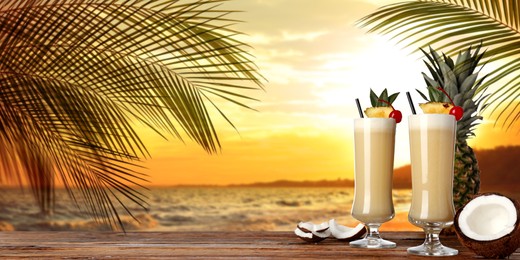 Image of Tasty Pina Colada cocktail on wooden table near ocean at sunset, space for text. Banner design