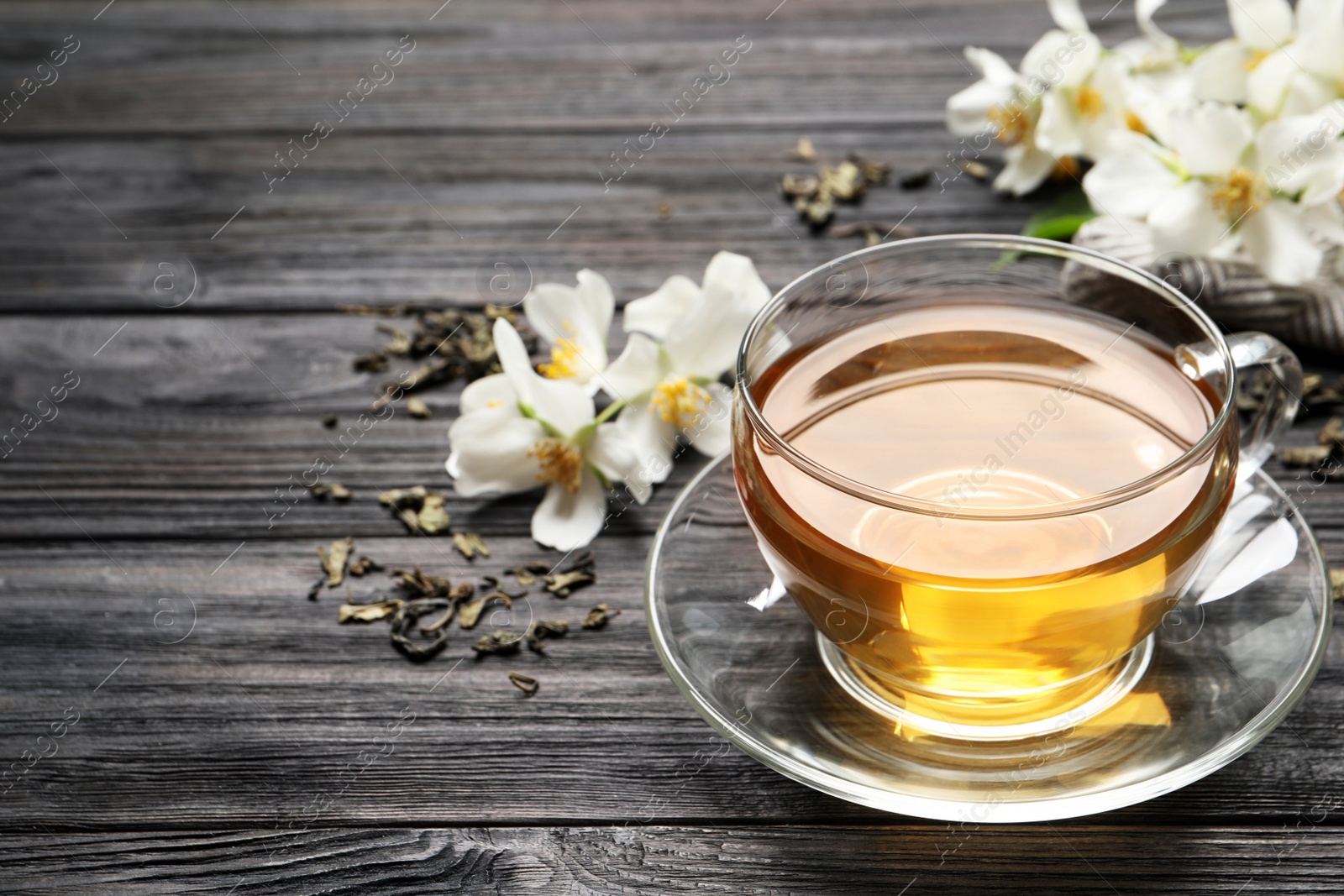 Photo of Glass cup of aromatic jasmine tea and fresh flowers on black wooden table, space for text