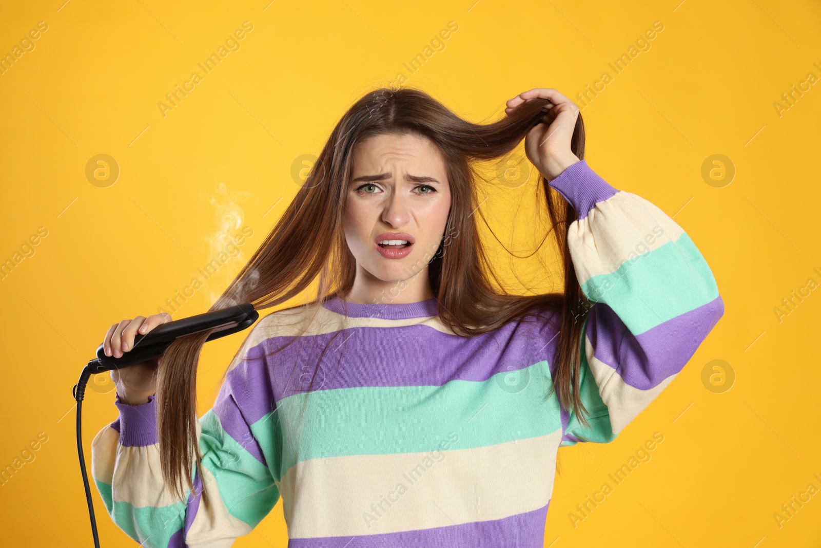 Photo of Stressed young woman with flattening iron on yellow background. Hair damage