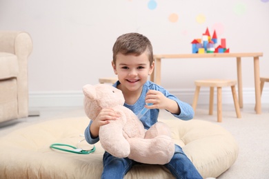 Photo of Cute child playing doctor with stuffed toy on floor in hospital