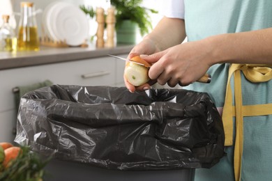 Woman peeling fresh onion with knife above garbage bin indoors, closeup