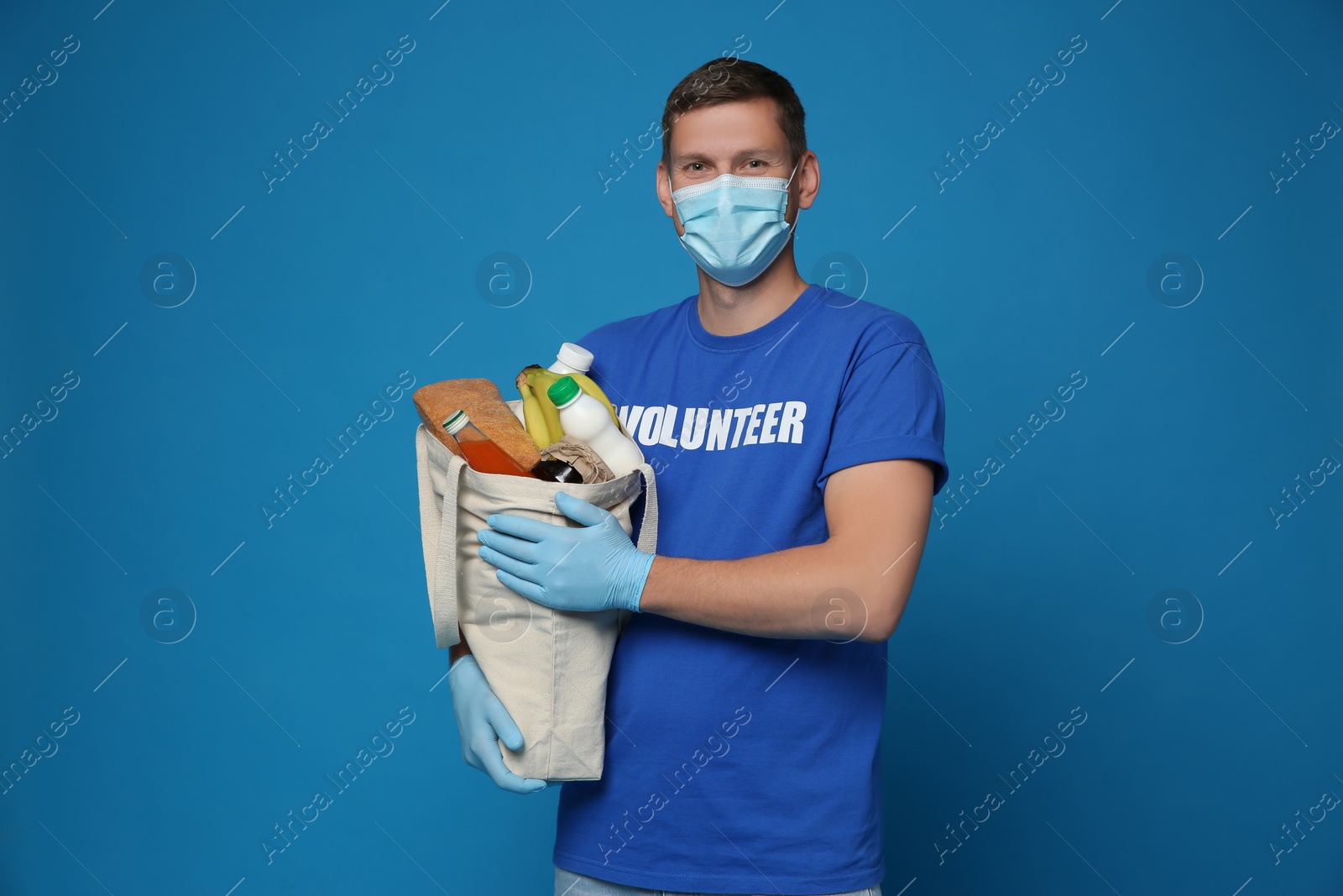 Photo of Male volunteer in protective mask and gloves with products on blue background. Aid during coronavirus quarantine	