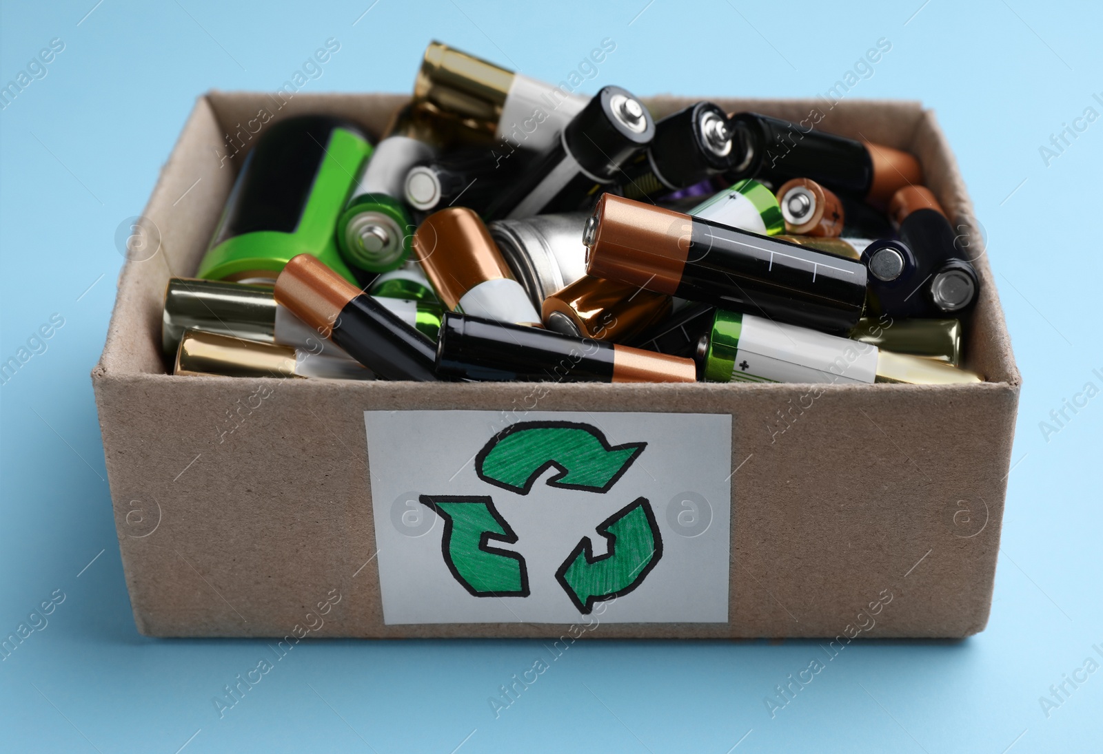 Image of Used batteries in cardboard box with recycling symbol on light blue background, closeup