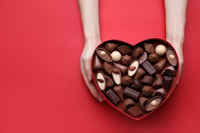 Woman holding heart shaped box with delicious chocolate candies on red background, top view. Space for text