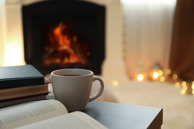 Photo of Cup of hot drink and books on table near fireplace at home. Cozy atmosphere