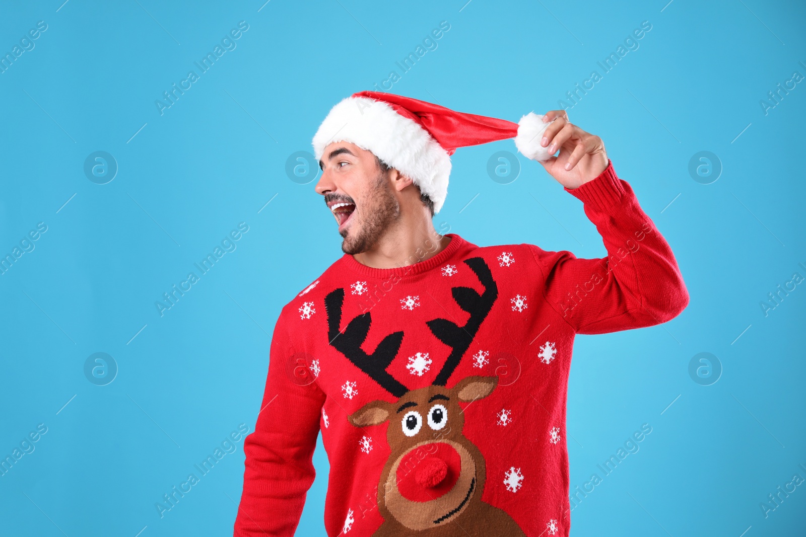 Photo of Emotional young man in Christmas sweater and Santa hat on light blue background