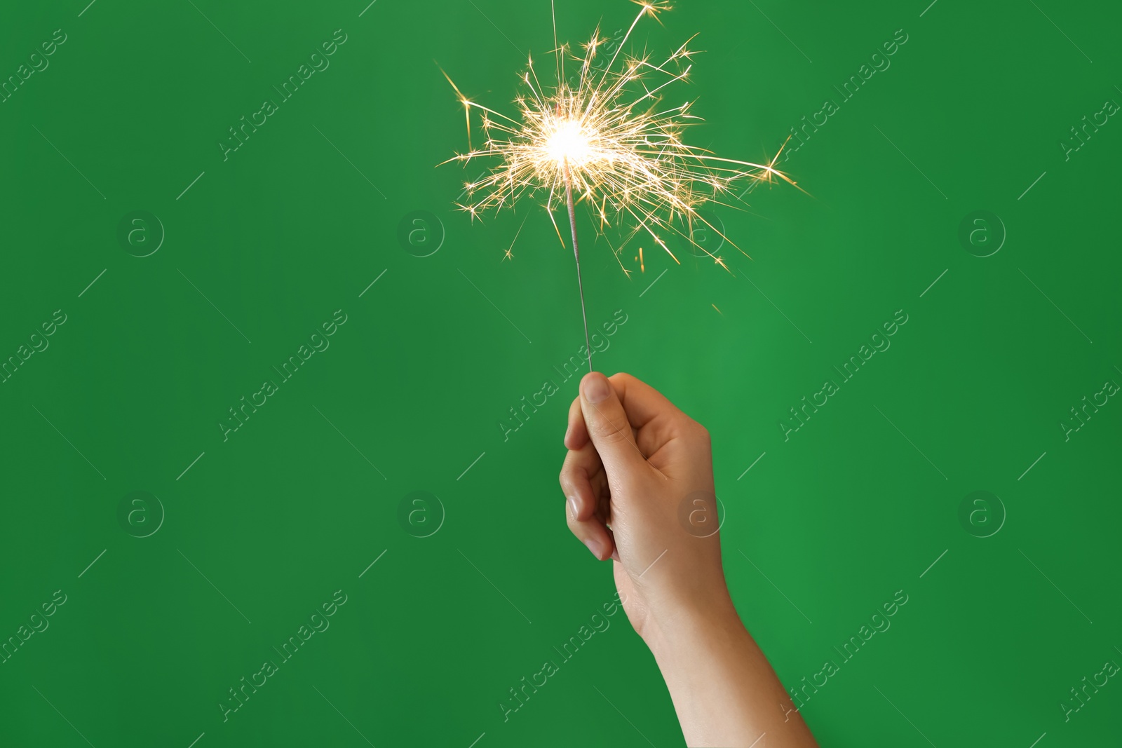 Photo of Woman holding bright burning sparkler on green background, closeup