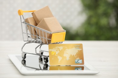 Online payment concept. Small shopping cart with bank card, boxes and tablet on white table, closeup