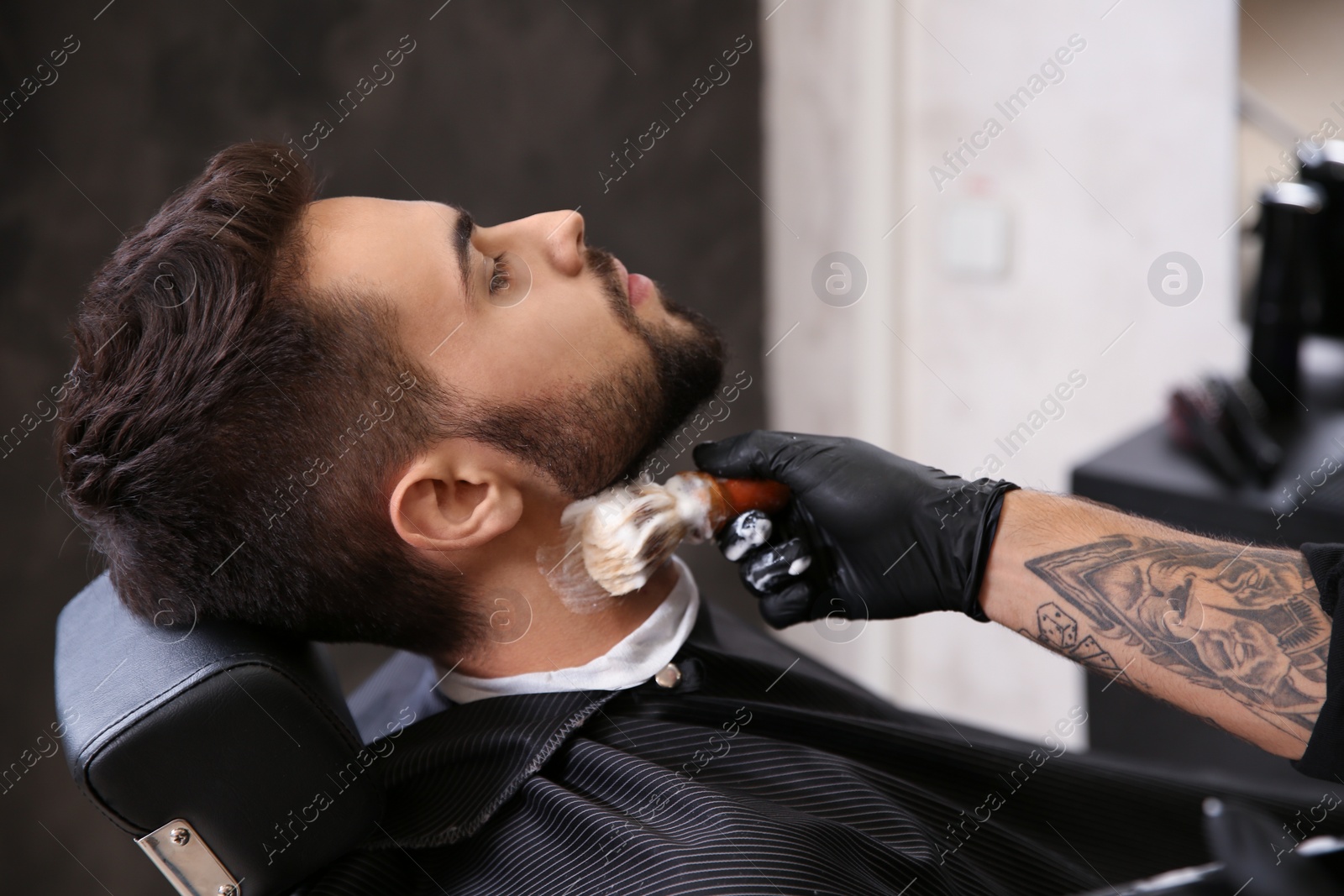 Photo of Professional hairdresser applying shaving foam onto client's skin in barbershop