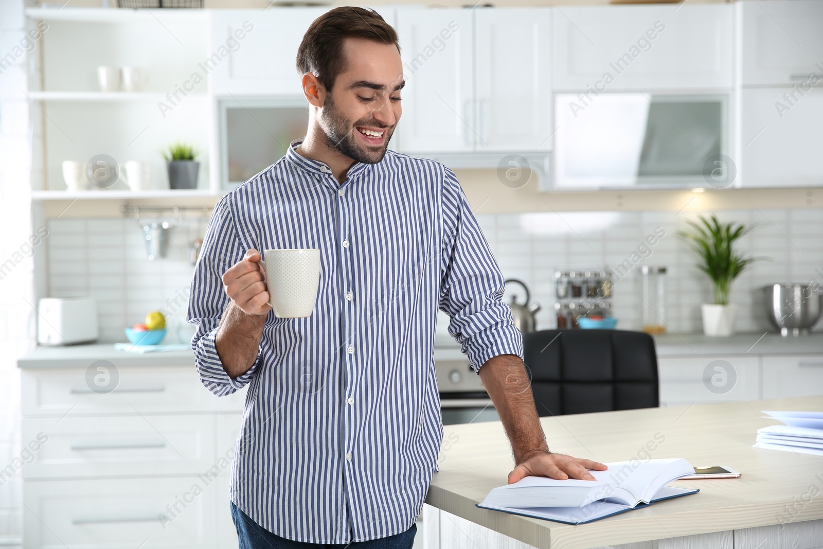 Photo of Handsome young man with cup of coffee reading book at home