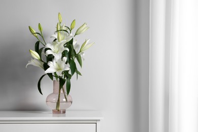 Beautiful bouquet of lily flowers in glass vase on table near white wall, space for text