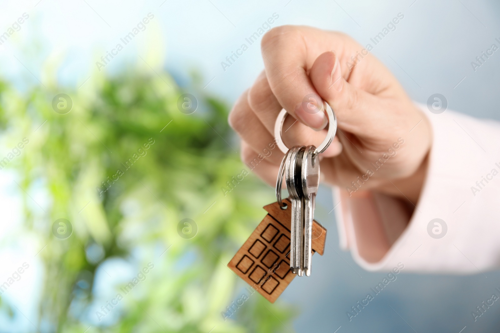 Photo of Woman holding house keys with trinket on blurred background, closeup. Space for text