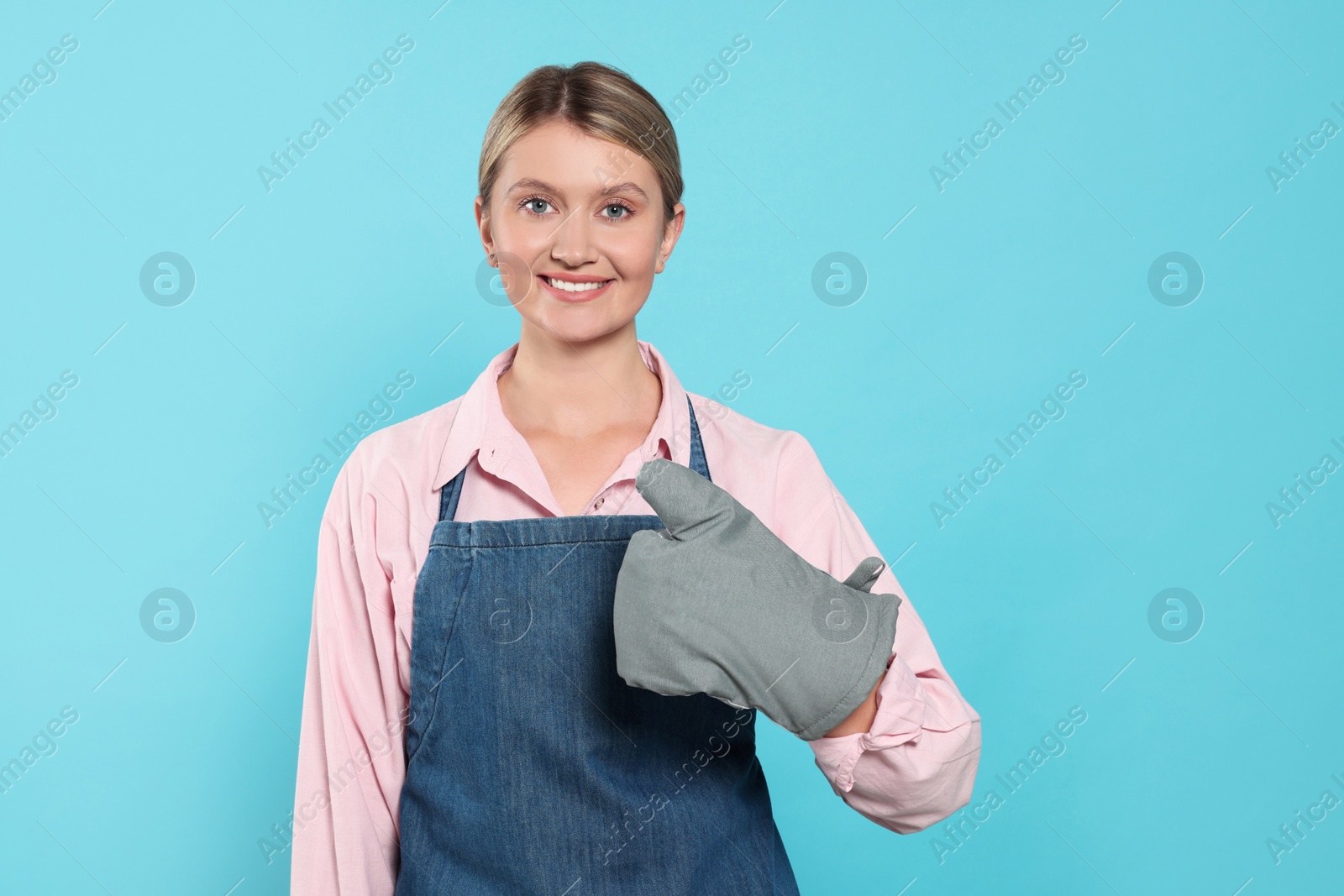 Photo of Beautiful young woman in denim apron and oven glove on light blue background