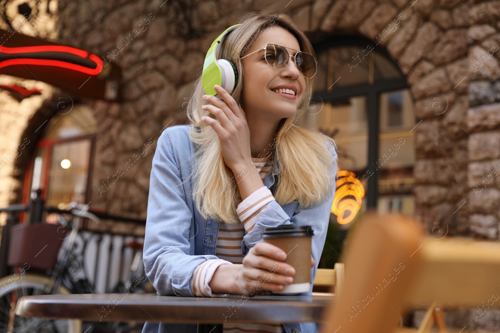 Photo of Happy young woman with coffee and headphones listening to music in outdoor cafe