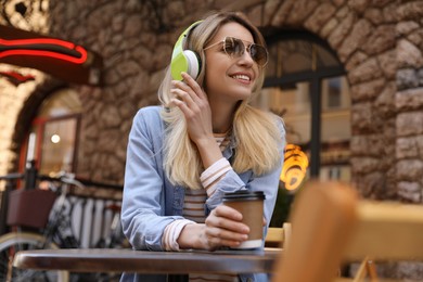 Photo of Happy young woman with coffee and headphones listening to music in outdoor cafe