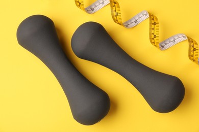 Dumbbells and measuring tape on yellow background, flat lay