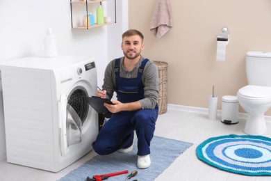 Young plumber with clipboard near washing machine in bathroom