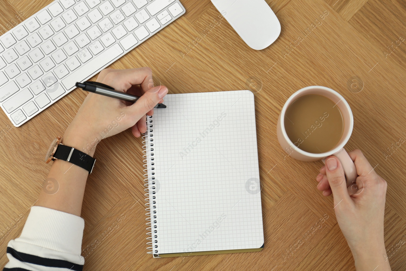 Photo of Left-handed woman with cup of coffee writing in notebook at wooden table, top view