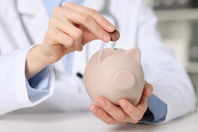 Photo of Doctor putting coin into piggy bank at white table indoors, closeup