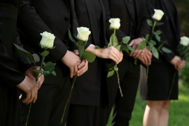 Photo of People in black clothes with white rose flowers outdoors, closeup. Funeral ceremony