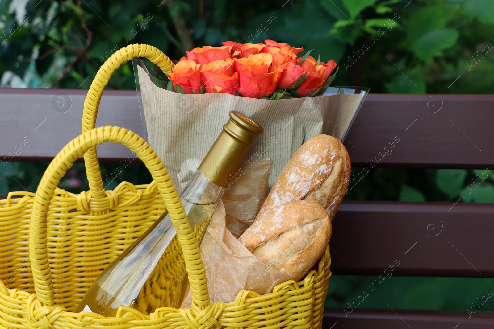 Photo of Yellow wicker bag with beautiful roses, bottle of wine and baguettes on bench outdoors