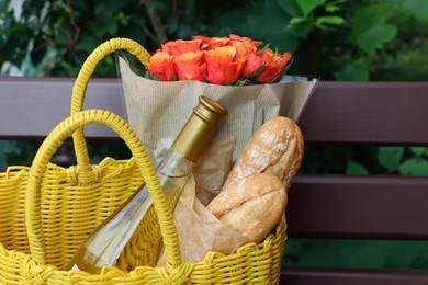 Yellow wicker bag with beautiful roses, bottle of wine and baguettes on bench outdoors