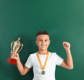 Happy boy with golden winning cup and medal near chalkboard