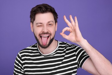 Photo of Happy man showing his tongue and making ok gesture on purple background