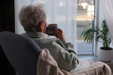 Photo of Elderly woman with cup of drink sitting in armchair indoors, space for text. Loneliness concept