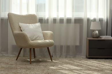 Photo of Soft white pillow on armchair and bedside table near window indoors