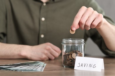 Photo of Financial savings. Man putting coin into glass jar at wooden table, closeup