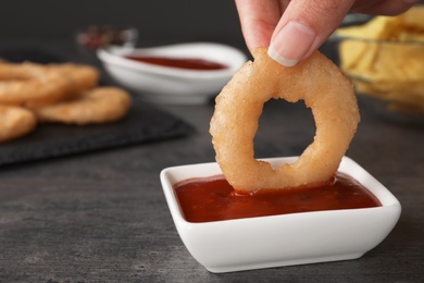 Woman dipping onion ring into hot chili sauce in bowl on table