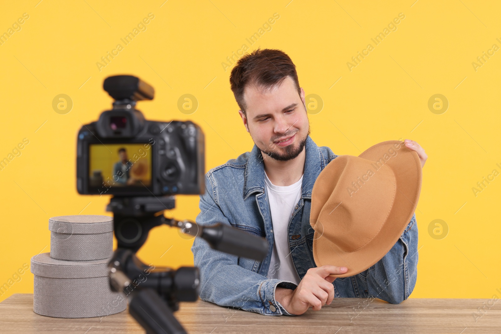 Photo of Smiling fashion blogger showing hat while recording video at table against orange background