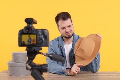 Smiling fashion blogger showing hat while recording video at table against orange background