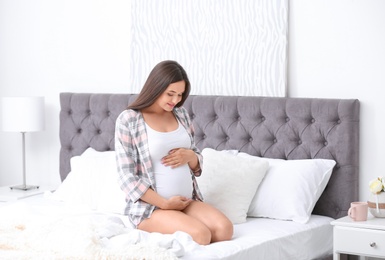 Photo of Young beautiful pregnant woman sitting on bed and touching her belly at home