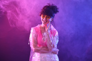 Photo of Beautiful young woman in transparent coat and sunglasses posing on color background in neon lights and smoke