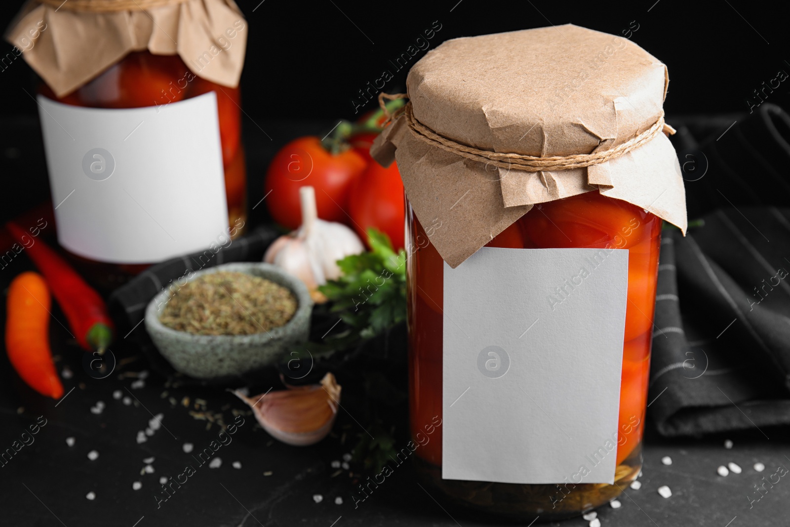 Photo of Jars of pickled tomatoes with blank stickers and products on black table