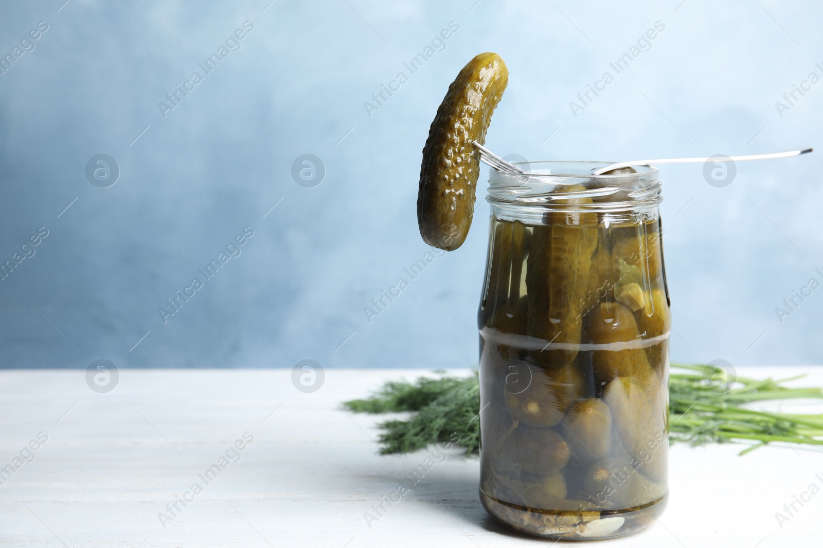 Photo of Fork with marinated cucumber over jar on white wooden table against blue background, space for text