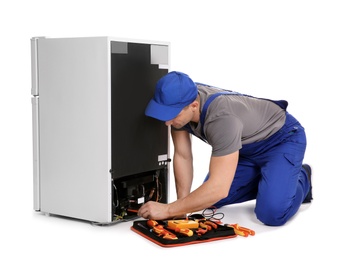 Male technician in uniform repairing refrigerator on white background