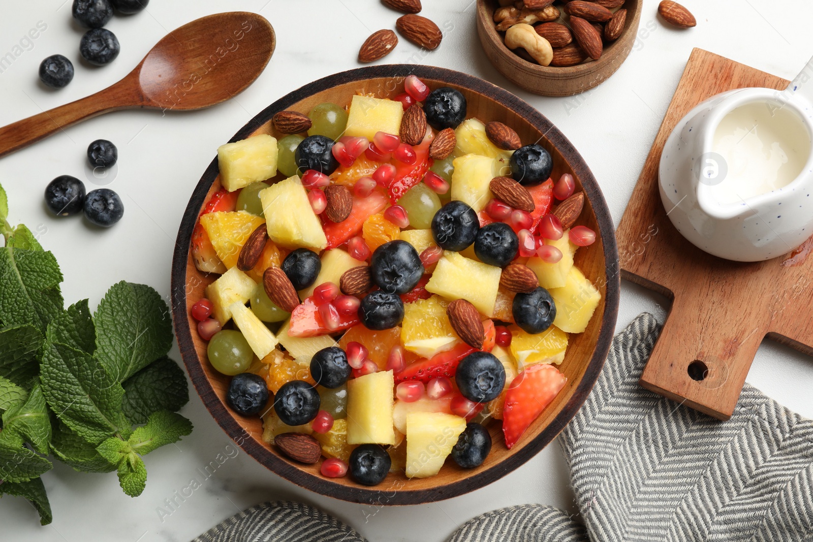 Photo of Delicious fruit salad in bowl, berries, fresh mint and nuts on white marble table, flat lay